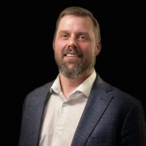 Smiling man with light brown hair and beard, wearing a navy blazer and white shirt, against a black background.
