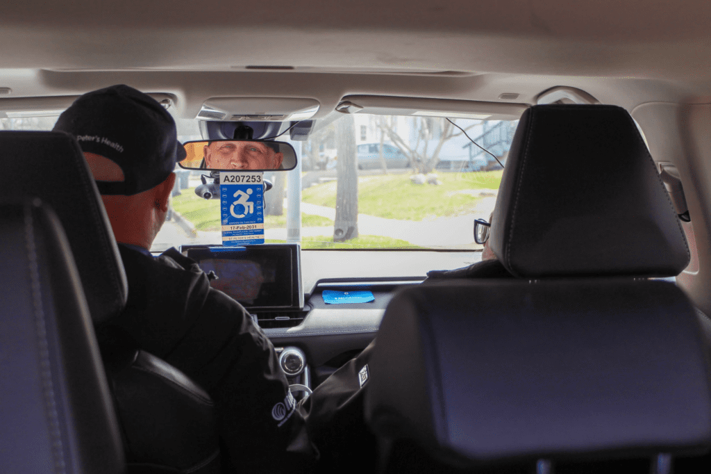 Interior view of a car with two people in the front seats; a blue handicap parking permit dangles from the rearview mirror.Outside street visible through the windshield.