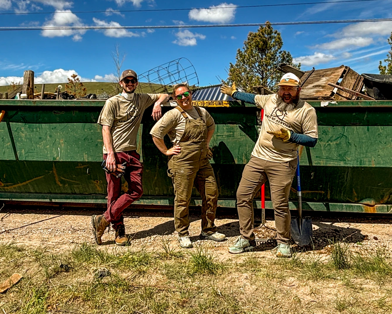 20260515-Williams-6 - Helena Area Chamber of Commerce Three volunteers in tan work clothes pose with shovels in front of a large green dumpster filled with debris under a sunny blue sky.