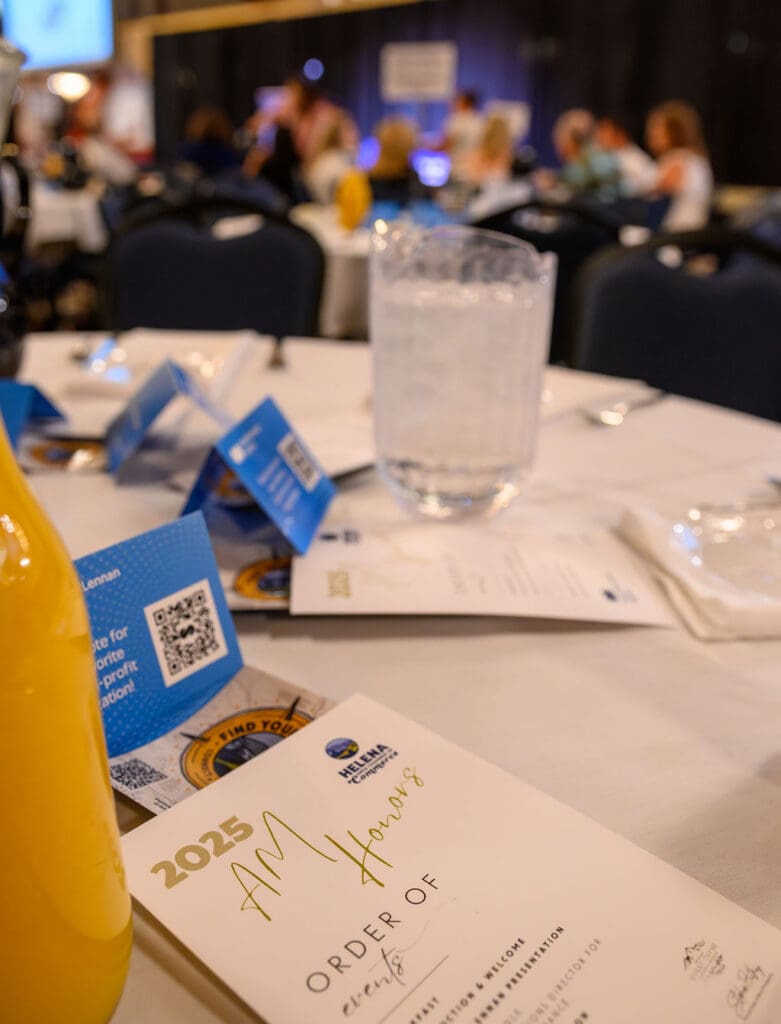 Close-up of a banquet table with a water pitcher, yellow drink bottle, blue event cards, and a '2025 Annual Honors' program on the table.