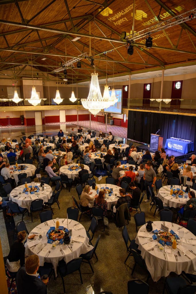 Banquet hall with round tables set for a formal dinner, chandeliers overhead, and a stage with podium and large screen in the background.