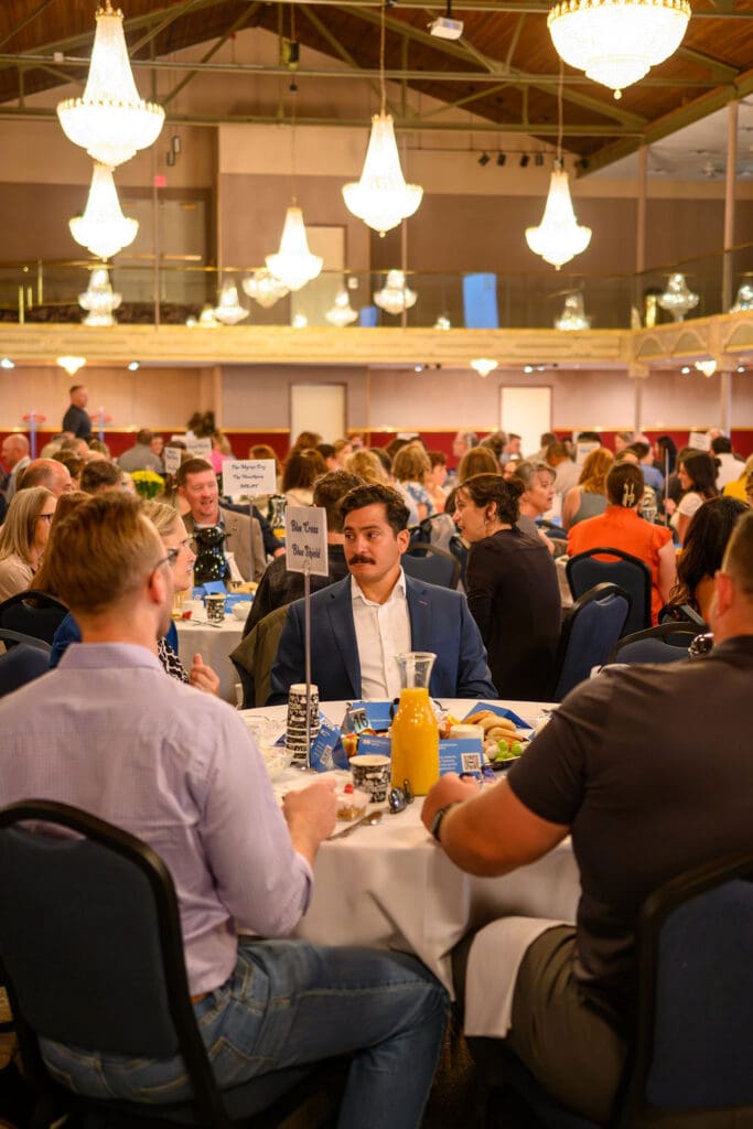 A large banquet hall filled with round tables and guests in business attire; foreground shows a man in a blue suit seated at a table.