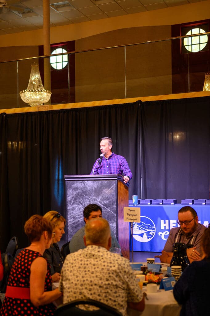 Man in a purple plaid shirt speaks at a wooden podium on a stage during a banquet or conference.