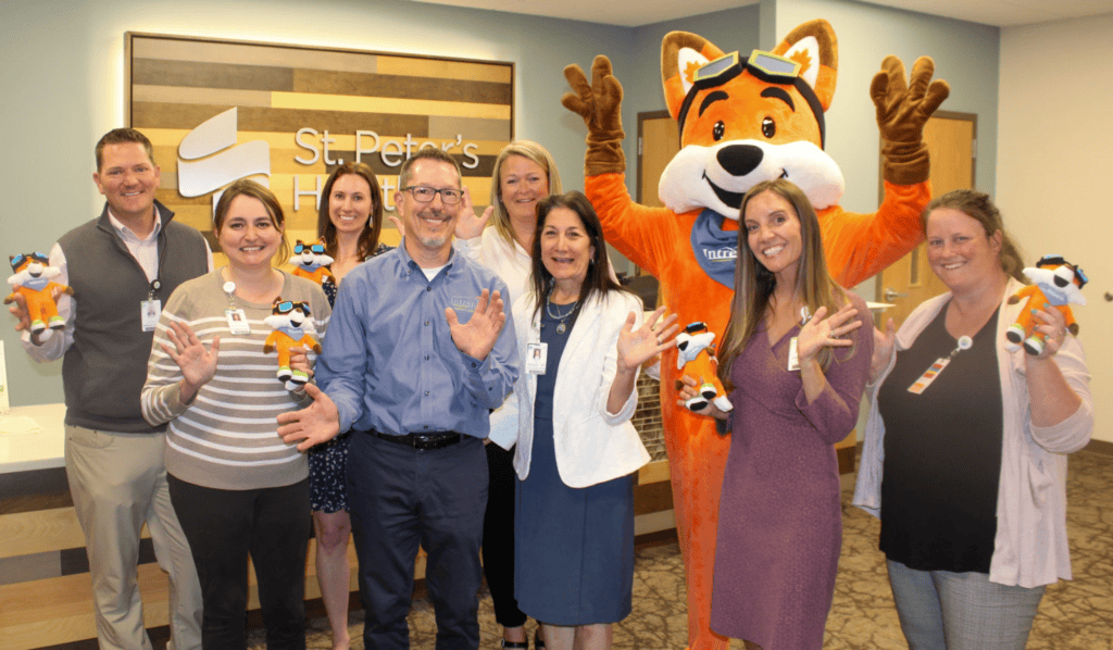 IMG_5015 - Helena Area Chamber of Commerce Group of hospital staff posing with a large orange fox mascot raising hands in celebration, in front of a St. Peter's Hospital sign.