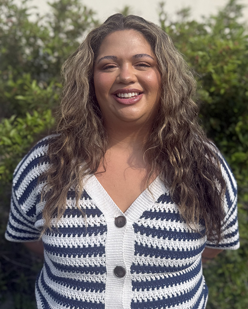 Allie Website - Helena Area Chamber of Commerce Smiling woman with long wavy hair standing outdoors in front of greenery, wearing a navy and white striped cardigan.
