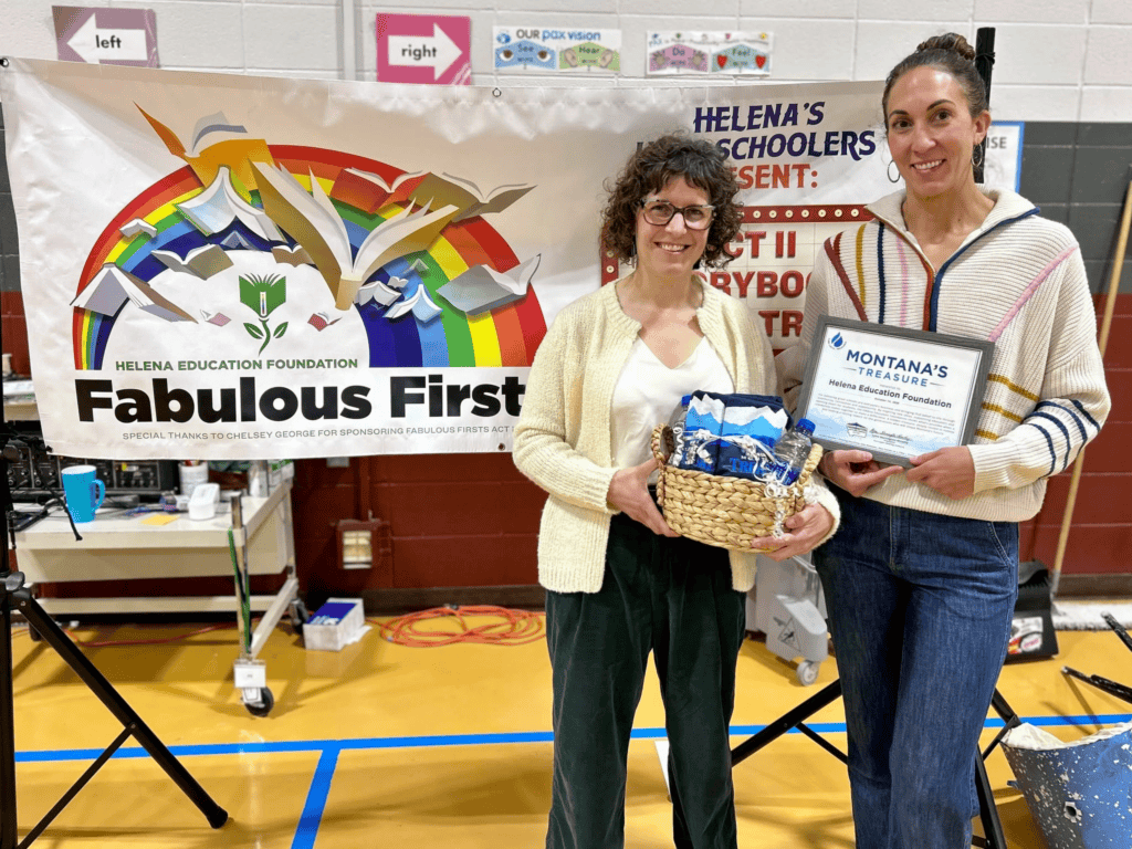 Two women stand in a gym next to a colorful banner reading 'Fabulous Firsts' and 'Helena Education Foundation' while one holds a trophy and gift basket and the other a framed certificate.